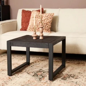 De Bijzettafel Boaz Zwart (60 cm) staat op een gedessineerd vloerkleed voor een beige bank met twee sierkussens en twee houten kandelaars, tegen een gedempte bruine muur.