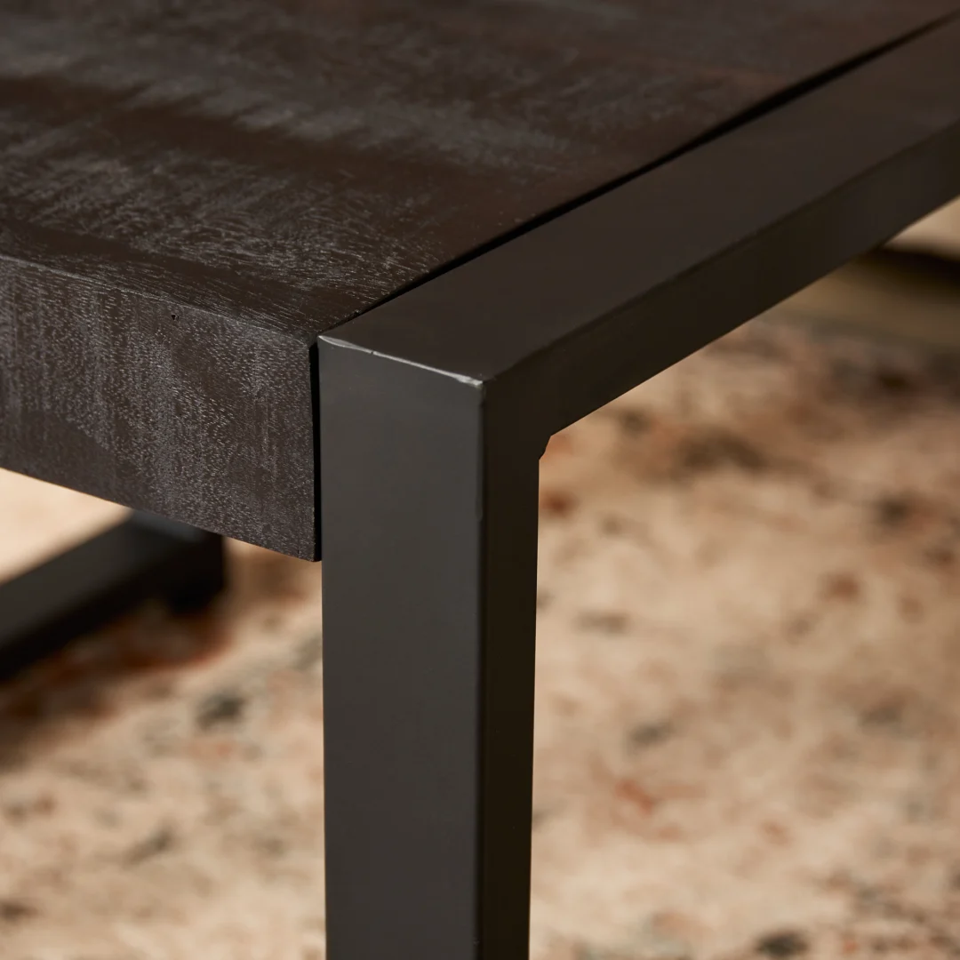 Close-up van de hoek van de Bijzettafel Boaz Zwart (60 cm) met een donker houten blad en zwart metalen frame, geplaatst op een beige tapijt met subtiele gespikkelde patronen.