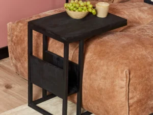 De Schuifregelaar Zwart mangohout, een klein zwart bijzettafeltje met een modern metalen frame, staat bij een bruine bank. Er liggen groene druiven en een beige beker op, tegen een roze muur en houten vloer.
