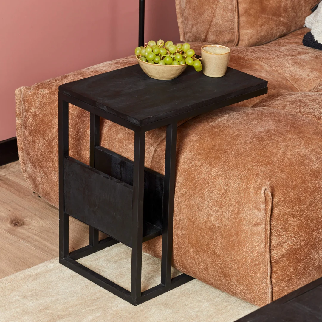 De Schuifregelaar Zwart mangohout, een klein zwart bijzettafeltje met een modern metalen frame, staat bij een bruine bank. Er liggen groene druiven en een beige beker op, tegen een roze muur en houten vloer.