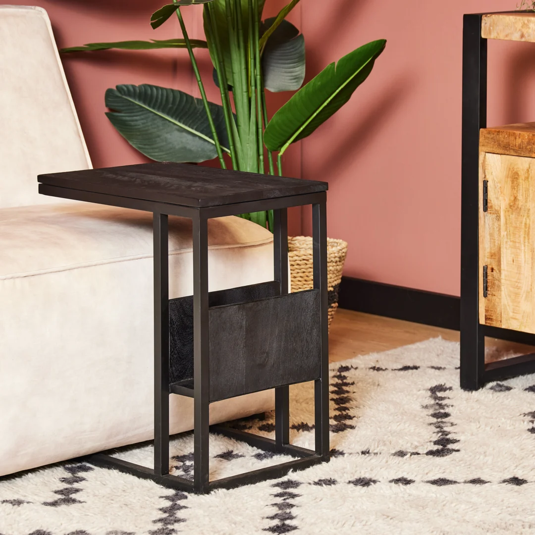 De Schuifregelaar Zwart mangohout, een moderne zwarte bijzettafel met een C-vormig metalen frame, staat naast een beige bank in een kamer met een roze muur, een vloerkleed met patroon, een bladplant, een gevlochten mand en een houten kast.