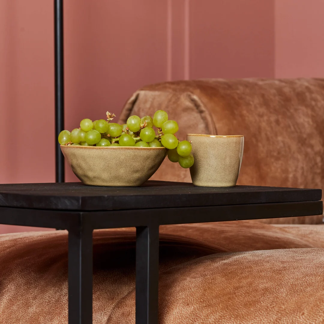 Een schaal met groene druiven en een beige beker rusten op de Schuifregelaar Zwart mangohout bijzettafel voor een bruin fluwelen bank, met een roze muur als achtergrond.