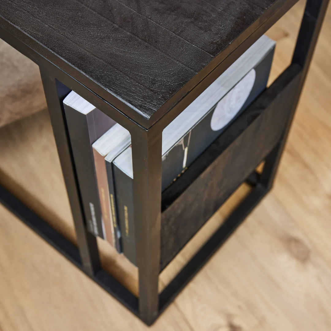 Een close-up van de Schuifregelaar Zwart mangohout bijzettafel met een ingebouwd tijdschriftenrek voor boeken en tijdschriften, op een lichte houten vloer.
