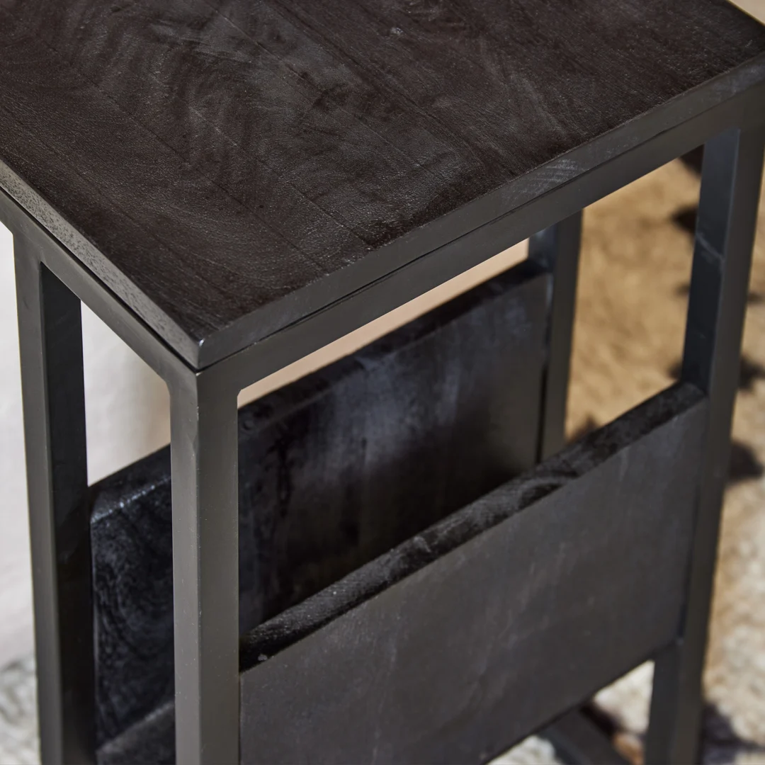 Een close-up van de Schuifregelaar bijzettafel van zwart mangohout, met een ingebouwde tijdschriftenhouder onder het tafelblad, op een licht tapijt met patroon.