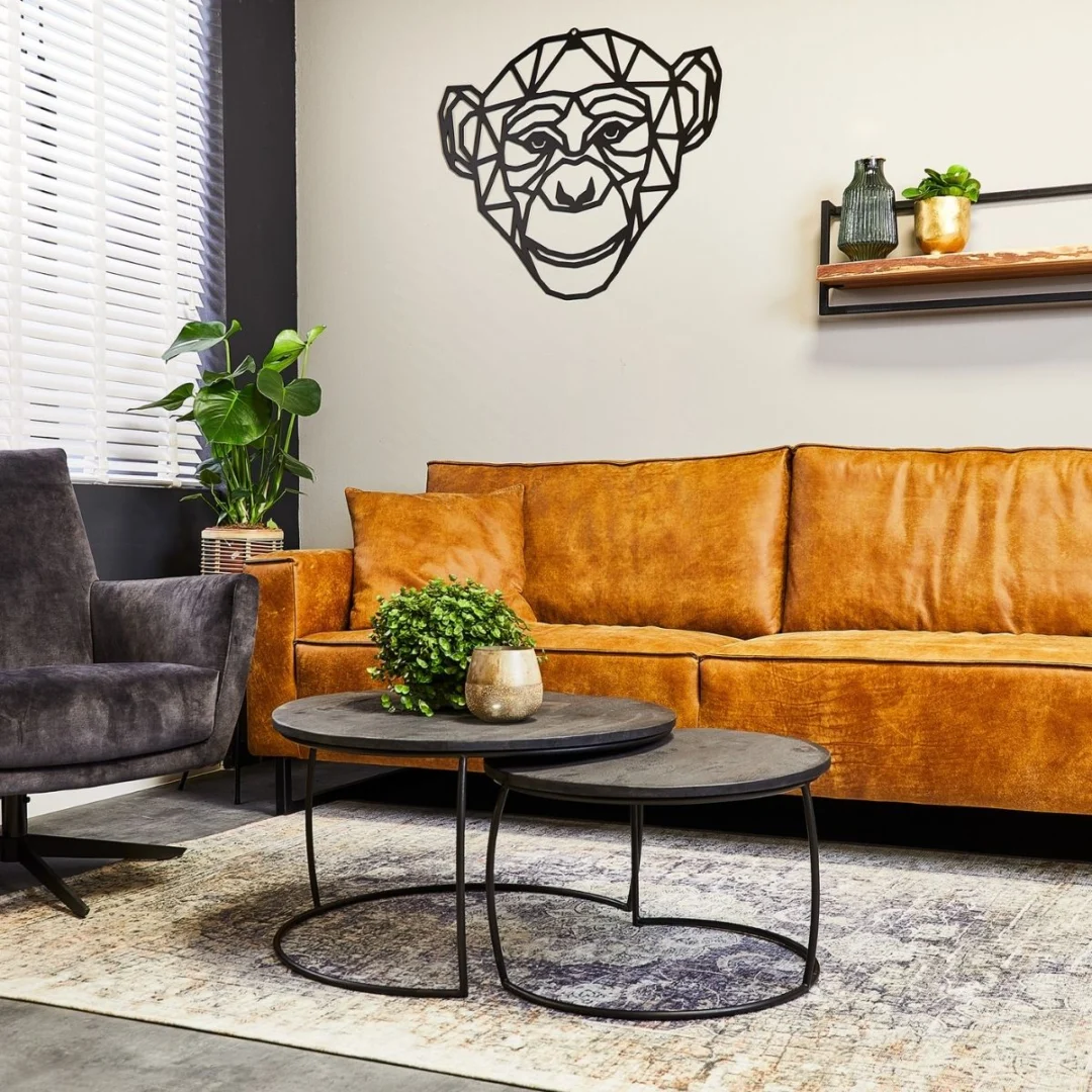 Een moderne woonkamer met een tan leren bank, grijze fauteuil, Salontafel Zwart 70 x 40 CM Rond als ronde zwarte salontafel met planten, een vloerkleed met patroon, geometrische metalen muurkunstwerk van een aap boven de bank en een decorplank aan de beige muur.