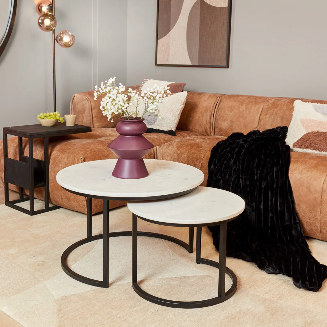 Een moderne woonkamer met een bruine leren bank, zwarte plaids en sierkussens. Aan de voorkant staan twee Salontafel Wit 70 x 50 CM Rond Marmer salontafels, met een paarse vaas met witte bloemen.