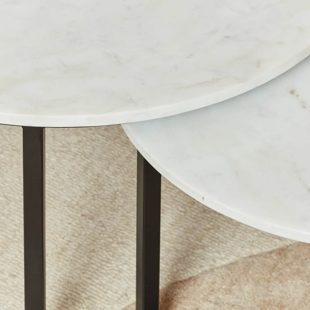 Close-up van twee Salontafels Wit 70 x 50 CM Rond Marmer met dunne zwarte metalen poten, naast elkaar geplaatst op een beige getextureerd tapijt.