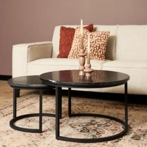 Een woonkamer met een crème bank, oranje en gedessineerde kussens en twee ronde zwart marmeren Salontafel Zwart nesttafels (70x70 cm). Op de grotere tafel staat een beige kandelaar met kaarsen en de vloer is bedekt met een vloerkleed met patroon.