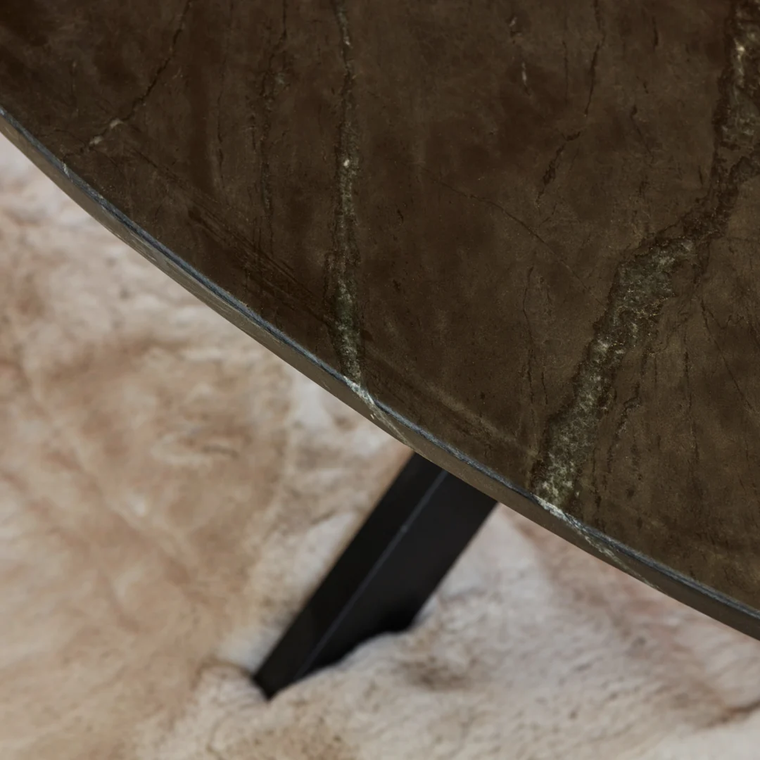 Close-up van de Salontafel Zwart 130 CM Ovaal Marmer: een ovaal, donker marmeren tafelblad met lichte adering, op zwart metalen poten en geplaatst op een zacht, beige vloerkleed.