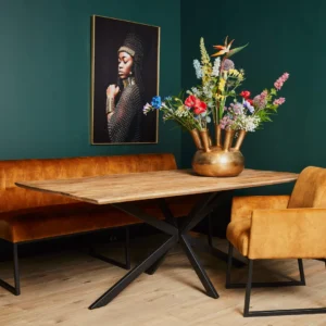 In een moderne eethoek staan de Eettafel Arlington 200 cm, een mosterdkleurige fluwelen bank en stoel en een grote vaas met kleurrijke bloemen. Aan de donkergroene muur hangt een ingelijst portret van een vrouw in profiel.