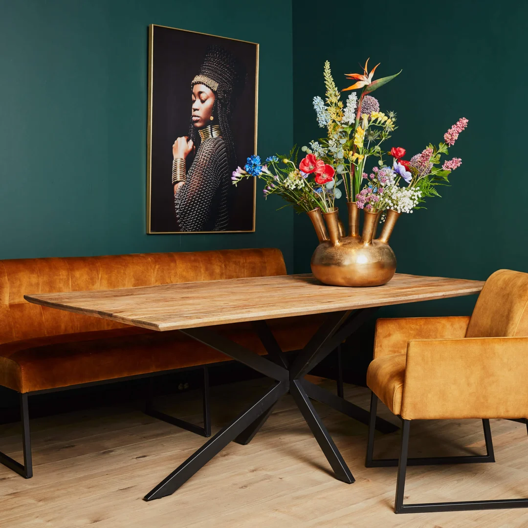 Eettafel Arlington met houten blad en metalen onderstel, gouden vaas met bloemen. Warme kleuren, zachte belichting, groene achtergrond.