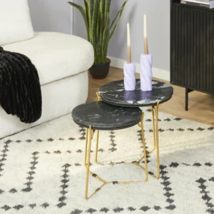 De Salontafel Zwart 45 x 45 CM Marmer, een ronde zwart marmeren nesttafel met gouden poten, staat op een gedessineerd vloerkleed. De kleinere tafel staat eronder en de set is vormgegeven met hoge kaarsen naast een witte bank en plant op de achtergrond.