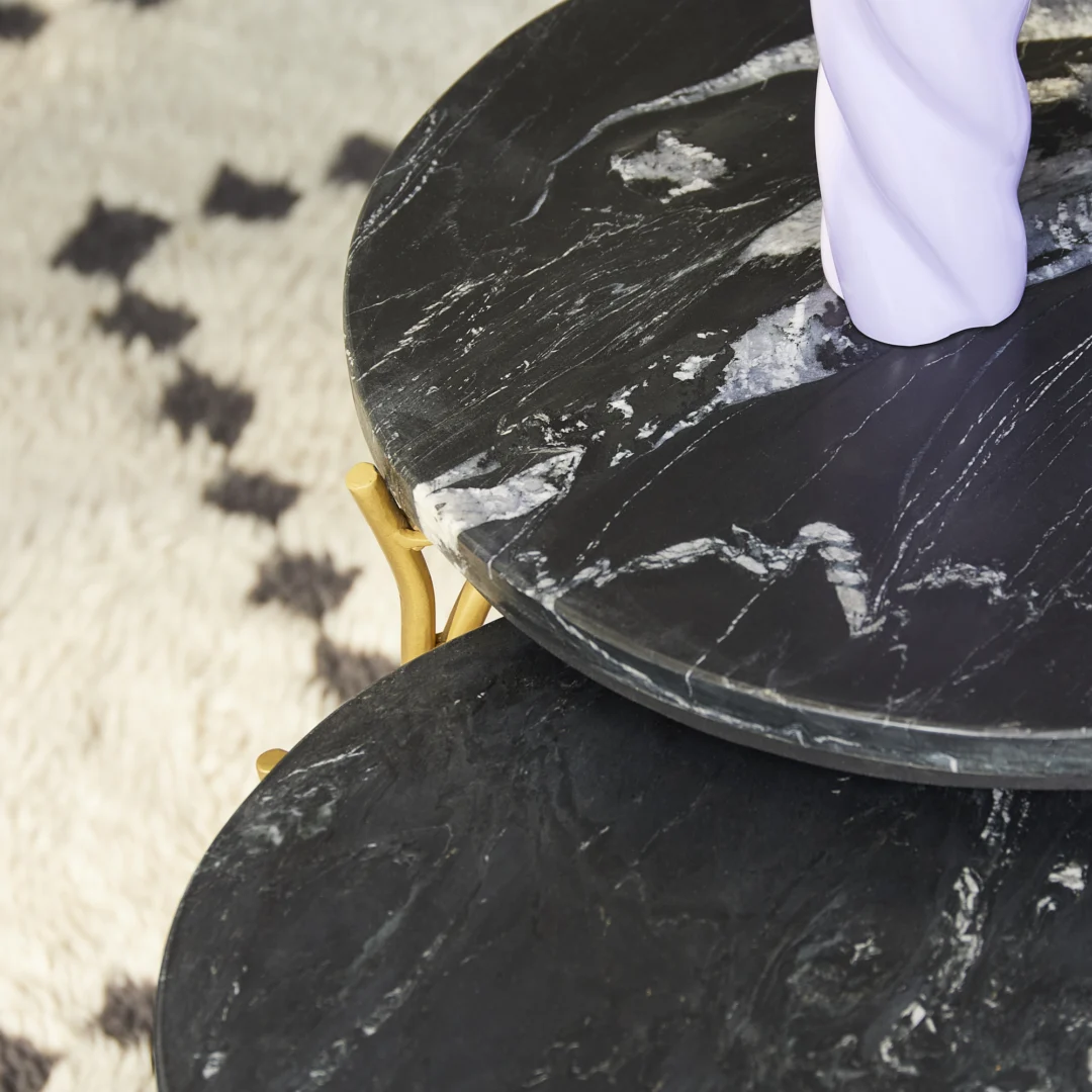 Close-up van de Salontafel Zwart 45 x 45 CM Marmer, met twee ronde zwart marmeren tafels met witte adering op verschillende hoogtes, goudkleurige metalen poten en een zwart-wit geometrisch tapijt met textuur op de achtergrond.
