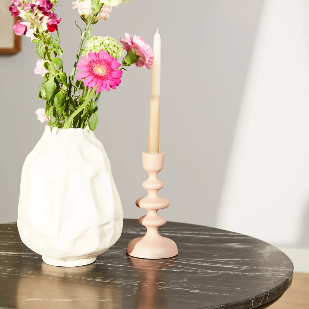 Een witte vaas met roze en witte bloemen en een roze kandelaar met een beige kaars staan op de Salontafel Zwart 75 x 75 CM Vierkant, een zwart marmeren salontafel, verlicht door zonlicht.