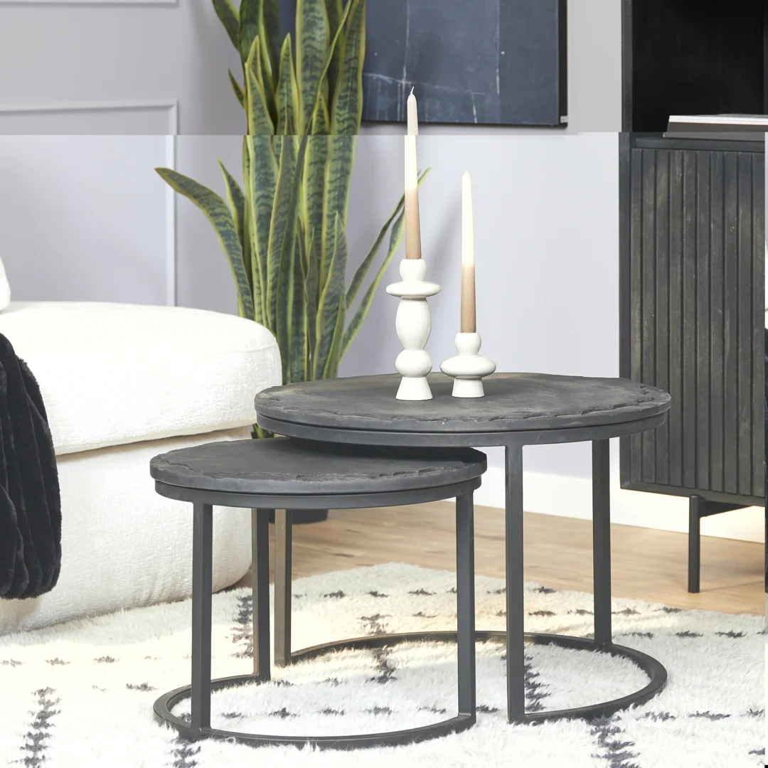 De Salontafel Zwart 61 x 61 CM Steen met slanke metalen poten staat op een vloerkleed met een licht patroon. Er staan twee witte kandelaars op met hoge kaarsen en op de achtergrond is een groene plant zichtbaar.