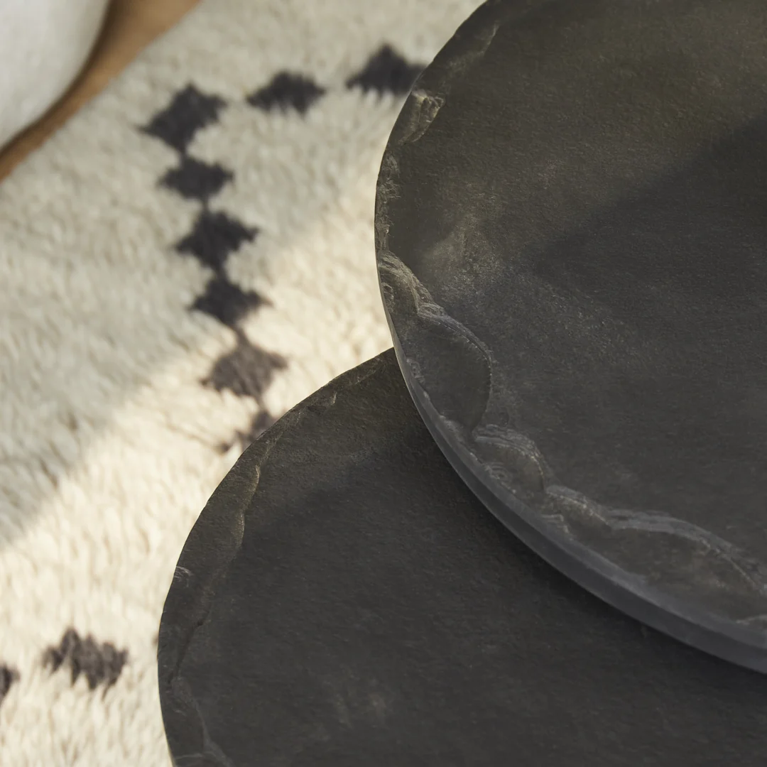 Close-up van twee gestapelde Salontafel Zwart 61 x 61 CM Steen tafels met onregelmatige randen, op een crèmekleurig tapijt met een zwart geometrisch ruitpatroon. Licht en schaduw voegen textuur toe aan de oppervlakken.