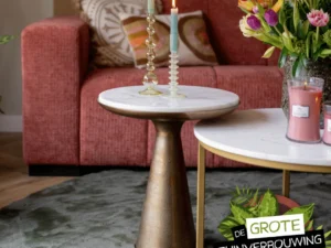 Een woonkamer met een rode bank, gedessineerde kussens, Bijzettafel Essy Large (wit marmer, 45 cm) als bijzettafel met twee brandende kaarsen en een vaas met kleurrijke bloemen. De tekst luidt: Gezien in De Grote Tuinverbouwing.