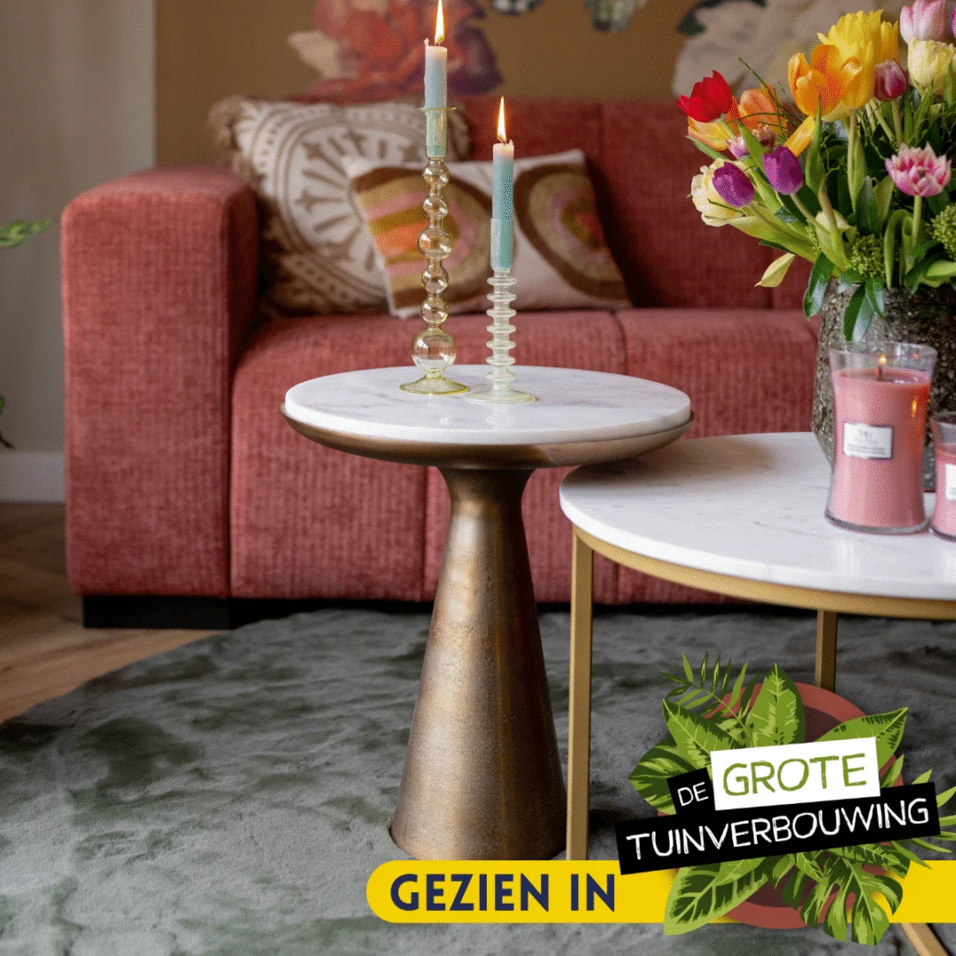 Een woonkamer met een rode bank, gedessineerde kussens, Bijzettafel Essy Large (wit marmer, 45 cm) als bijzettafel met twee brandende kaarsen en een vaas met kleurrijke bloemen. De tekst luidt: Gezien in De Grote Tuinverbouwing.