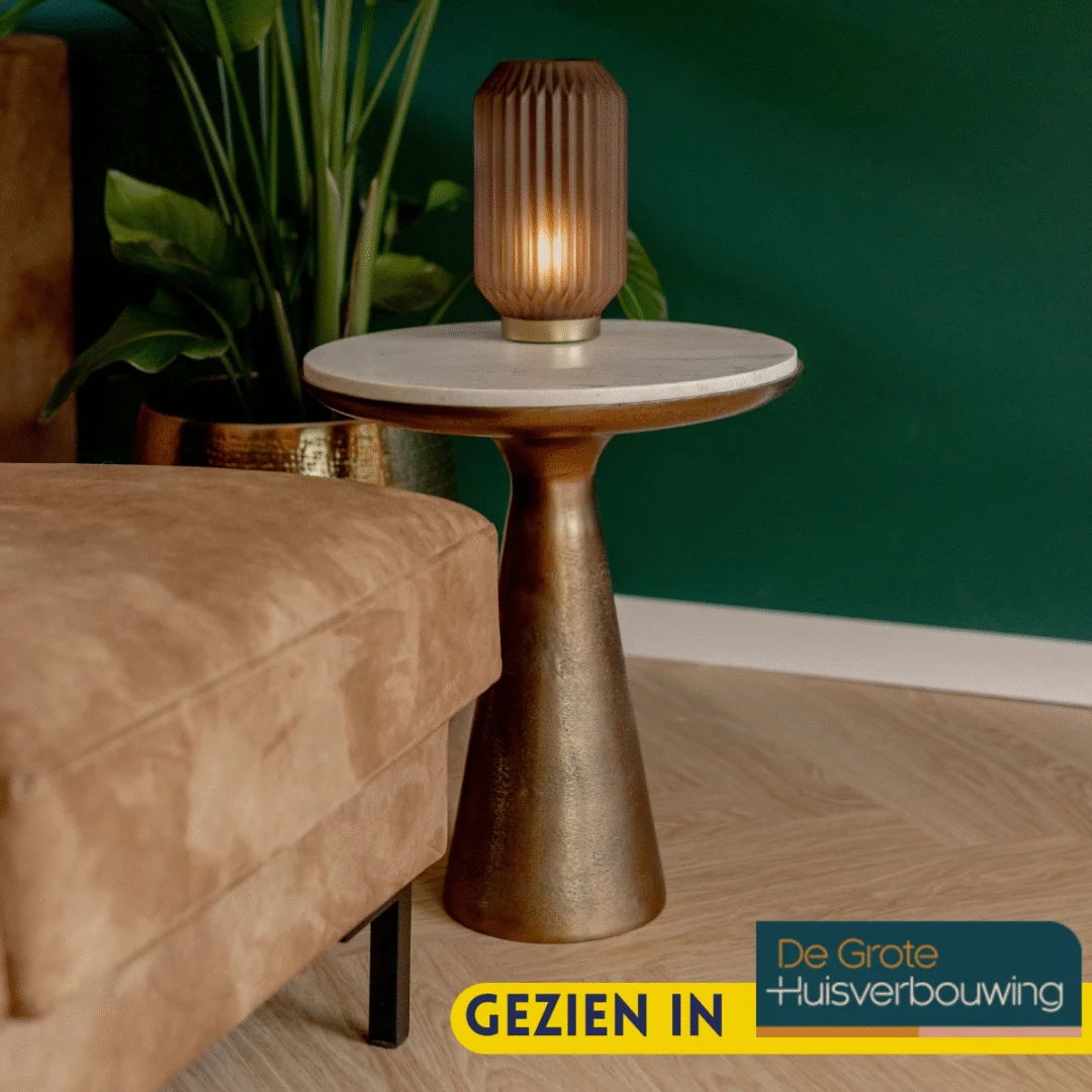 Een Bijzettafel Essy Large Wit marmer 45 cm met een gouden voet houdt een bruine geribbelde glazen lamp vast. Daarnaast staan een bruine bank en een groene plant. Op een label staat GEZIEN IN De Grote Huisverbouwing.