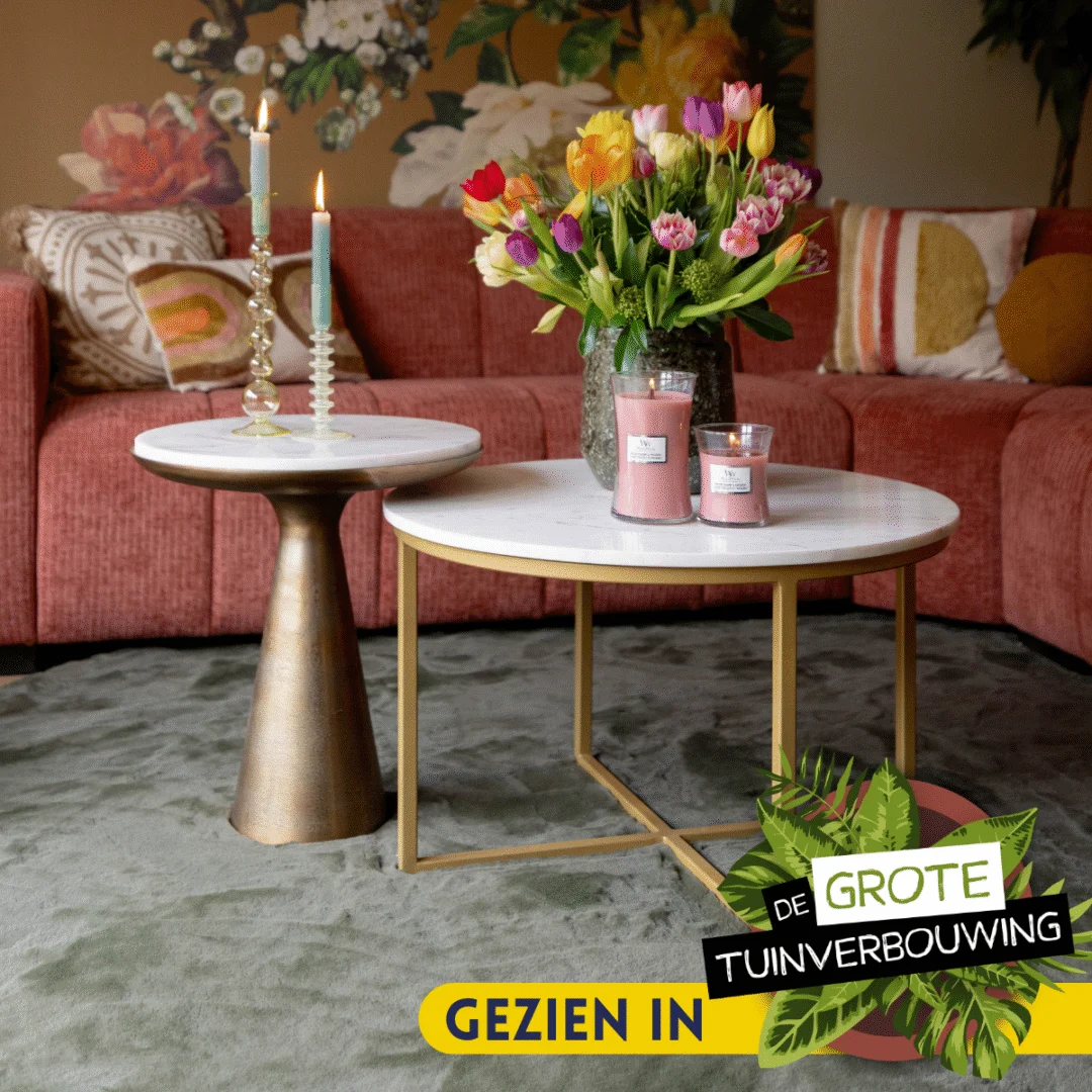 De Bijzettafel Essy Large | Wit marmer 45 cm is gestyled met kaarsen en een vaas met kleurrijke tulpen in een gezellige woonkamer met een roze bank, bloemen muurkunst en een groen vloerkleed. Tekst: GEZIEN IN DE GROTE TUINVERBOUWING.
