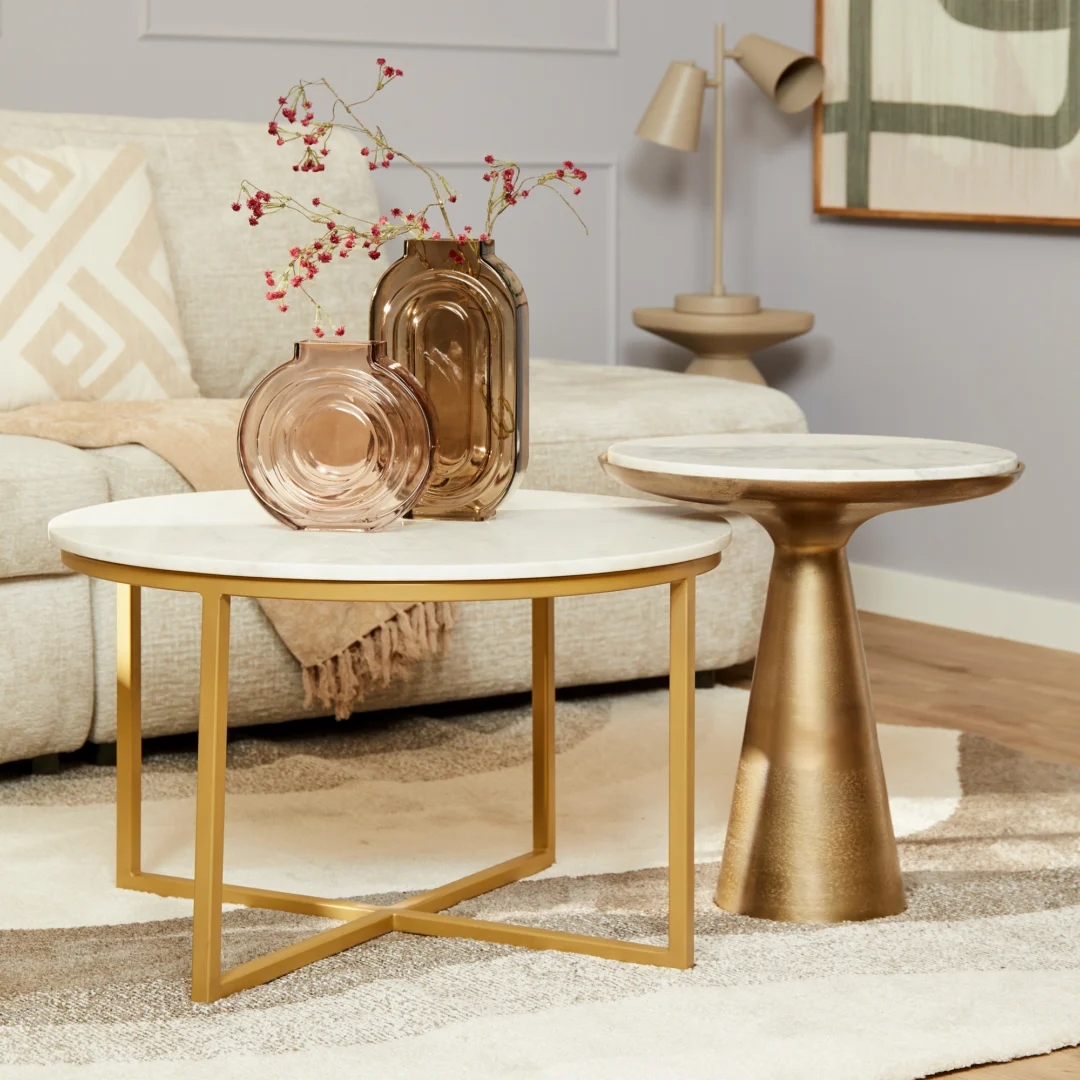 Een woonkamer met de Bijzettafel Essy Large (wit marmer, 45 cm) met gouden poten, een bijpassende gouden bijzettafel, decoratieve vazen met takken, een beige bank en een moderne neutrale inrichting.