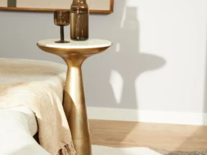 De Bijzettafel Essy Small Wit marmer 38 cm, met een gouden basis en wit marmeren blad, houdt een bruine glazen vaas en bokaal naast een beige bed met een crèmekleurig kleed in een zacht verlichte moderne kamer.