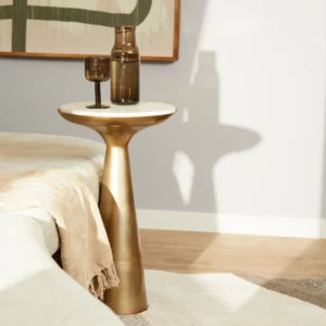De Bijzettafel Essy Small Wit marmer 38 cm, met een gouden basis en wit marmeren blad, houdt een bruine glazen vaas en bokaal naast een beige bed met een crèmekleurig kleed in een zacht verlichte moderne kamer.