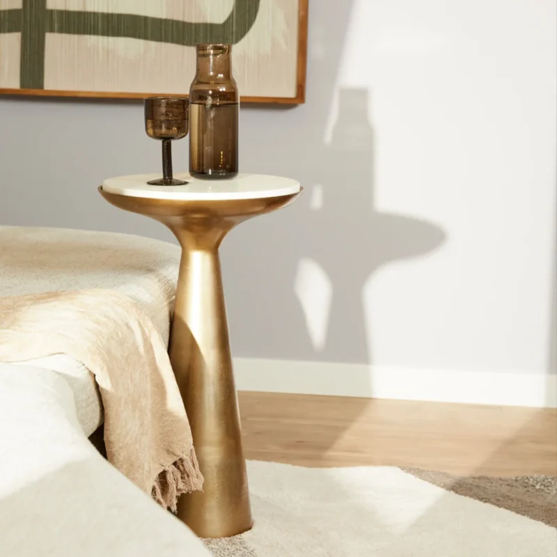 De Bijzettafel Essy Small Wit marmer 38 cm, met een gouden basis en wit marmeren blad, houdt een bruine glazen vaas en bokaal naast een beige bed met een crèmekleurig kleed in een zacht verlichte moderne kamer.