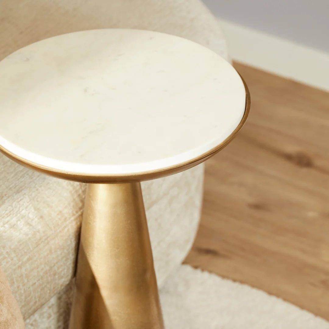 De Bijzettafel Essy Small met een wit marmeren blad van 38 cm staat op een gouden sokkel naast een beige bank op een houten vloer.