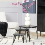 Een moderne woonkamer met een witte bank, zwarte plaids en drie geneste Salontafel Zwart 50 CM Marmer tafels. Op de grootste tafel staat een witte sculpturale vaas met felroze bloemen, met een zwarte kast en abstracte kunst op de achtergrond.
