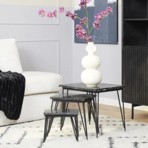 Een moderne woonkamer met een witte bank, zwarte plaids en drie geneste Salontafel Zwart 50 CM Marmer tafels. Op de grootste tafel staat een witte sculpturale vaas met felroze bloemen, met een zwarte kast en abstracte kunst op de achtergrond.