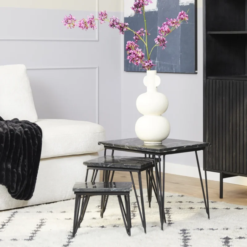Een moderne woonkamer met een witte bank, zwarte plaids en drie geneste Salontafel Zwart 50 CM Marmer tafels. Op de grootste tafel staat een witte sculpturale vaas met felroze bloemen, met een zwarte kast en abstracte kunst op de achtergrond.