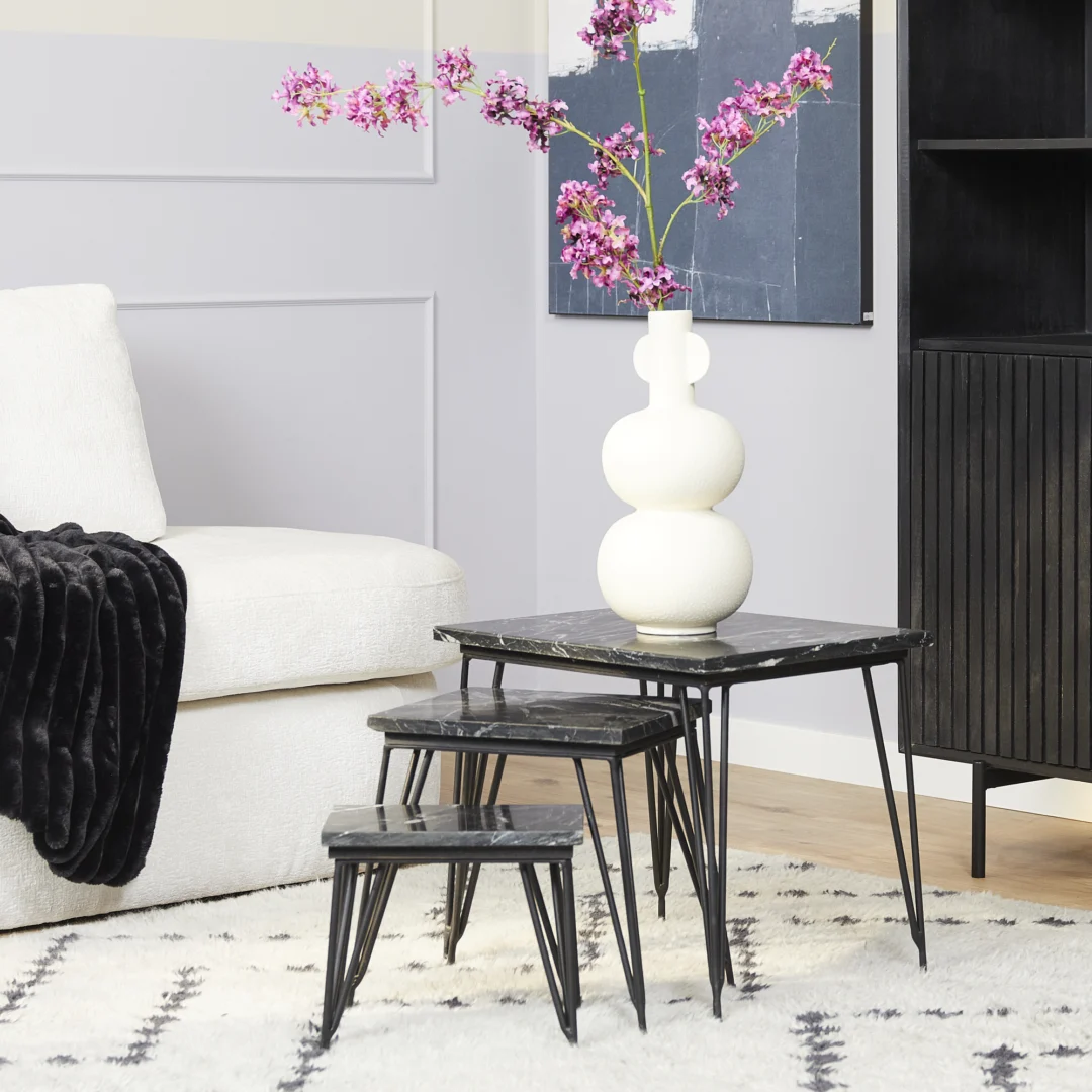 Een moderne woonkamer met een witte bank, zwarte plaids en drie geneste Salontafel Zwart 50 CM Marmer tafels. Op de grootste tafel staat een witte sculpturale vaas met felroze bloemen, met een zwarte kast en abstracte kunst op de achtergrond.