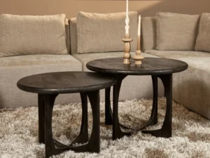 De Salontafel Milou | Zwart | 70 cm, een ronde salontafel van donker hout, staat op een beige shagtapijt voor een tan sofa en toont twee gebeeldhouwde houten kandelaars met witte kaarsen.