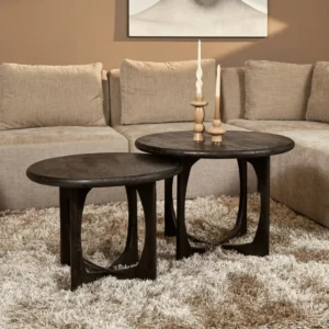 De Salontafel Milou | Zwart | 70 cm, een ronde salontafel van donker hout, staat op een beige shagtapijt voor een tan sofa en toont twee gebeeldhouwde houten kandelaars met witte kaarsen.