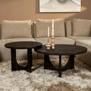 De Salontafel Zwart 60 CM, een ronde salontafel van donker hout met een geometrisch onderstel, staat op een beige shagtapijt voor een lichtbeige sectionaal. Op de tafel staan drie decoratieve kaarsen en aan de muur hangt een ingelijst kunstwerk.