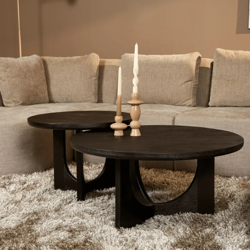 Een Salontafel Zwart 60 CM met gebogen poten staat op een beige shaggy vloerkleed voor een beige sectionaal, met daarop drie houten kandelaars met witte kaarsen.