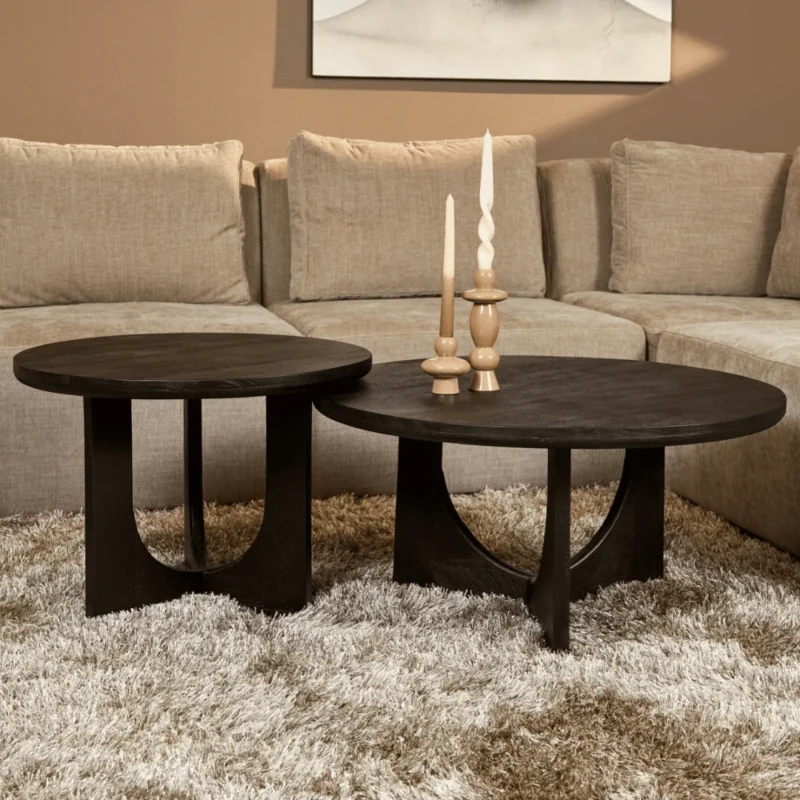 Een Salontafel Zwart 60 CM ligt op een pluche beige vloerkleed voor een tan sectional. Boven de tafel staan drie houten kandelaars met witte kaarsen en daarboven hangt een neutraal kunstwerk.