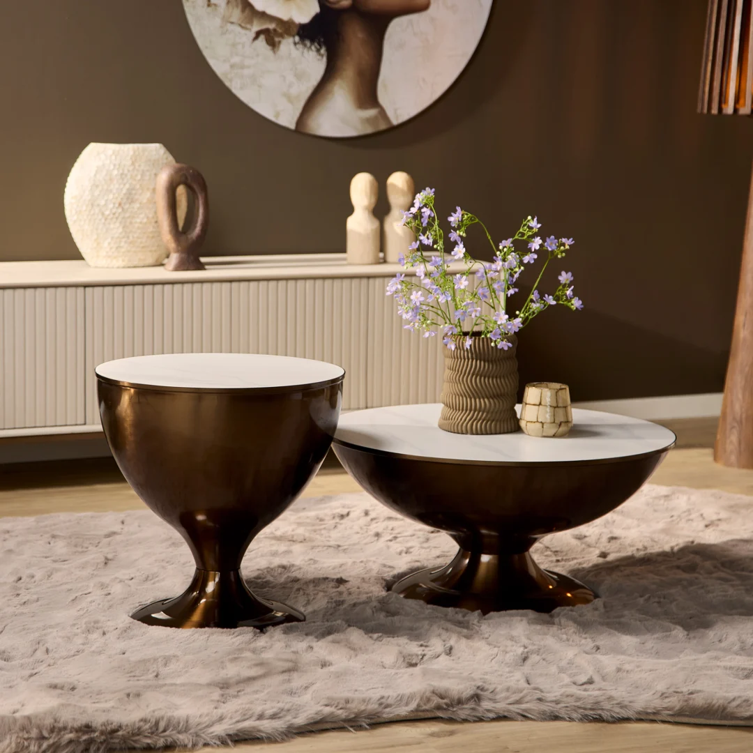 Twee Salontafel Opbergvak tafels met bronzen onderstellen en witte bladen rusten op een beige vloerkleed met textuur. Op één tafel staan een vaas met paarse bloemen en een klein beeldje, terwijl op de achtergrond decoratieve voorwerpen en kunst te zien zijn.