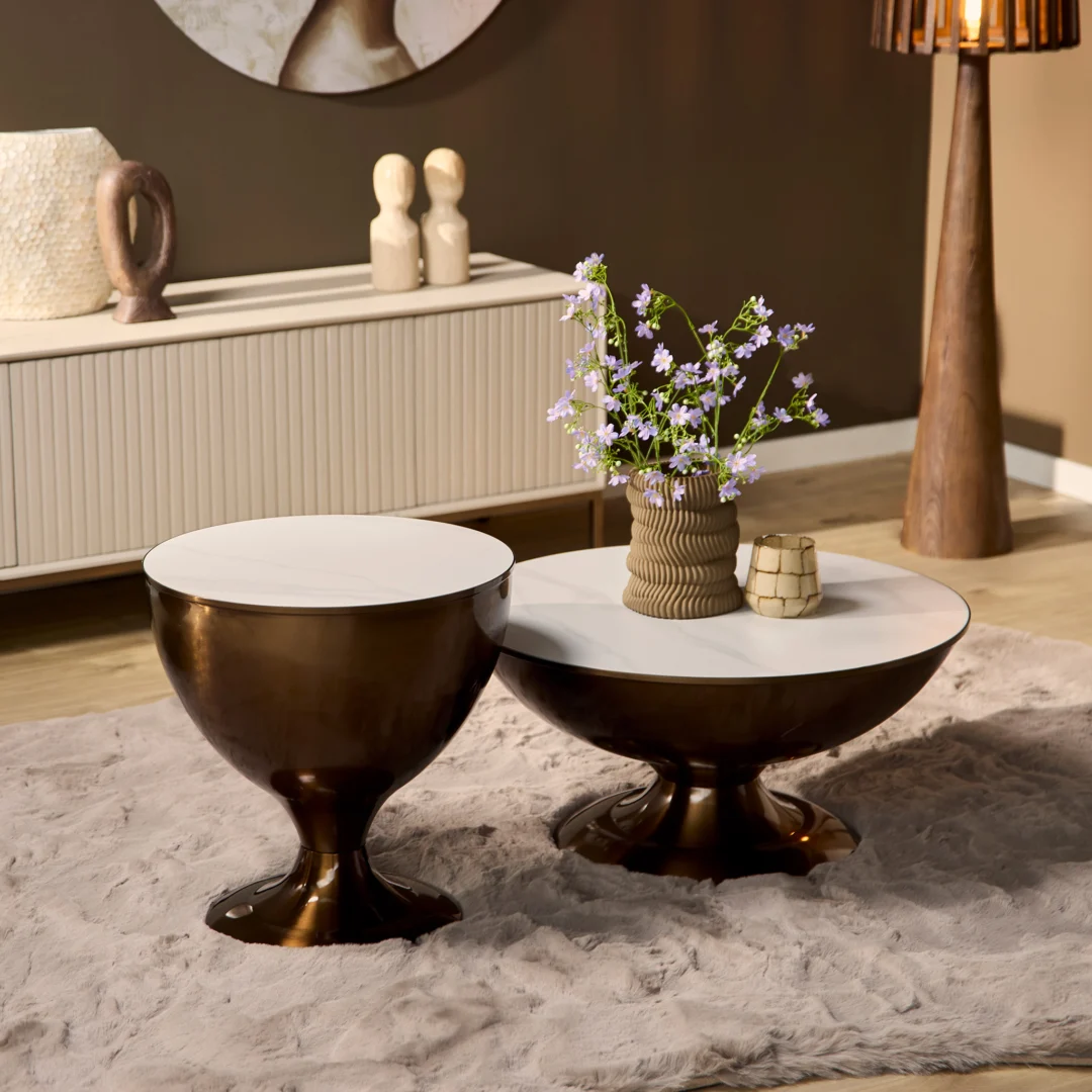 De Salontafel Opbergvak, met een modern rond ontwerp met een bronzen basis en wit blad, staat op een pluche beige vloerkleed. Een vaas met paarse bloemen en een klein decorstuk staan erop, met een neutrale inrichting en warme verlichting op de achtergrond.
