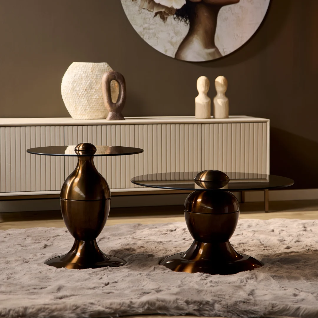 Een paar Salontafel Industrieel bronzen bijzettafeltjes met ronde glazen bladen staan op een beige vloerkleed voor een wit dressoir, geaccentueerd door vazen en gedeeltelijke portretkunst op de achtergrond.