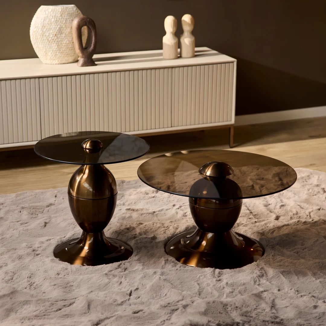 Twee Salontafel Industrieel bronzen bijzettafels met ronde, getinte glazen bladen rusten op een beige getextureerd tapijt in een minimalistische woonkamer; een bleke console en abstract decor zijn te zien op de achtergrond.