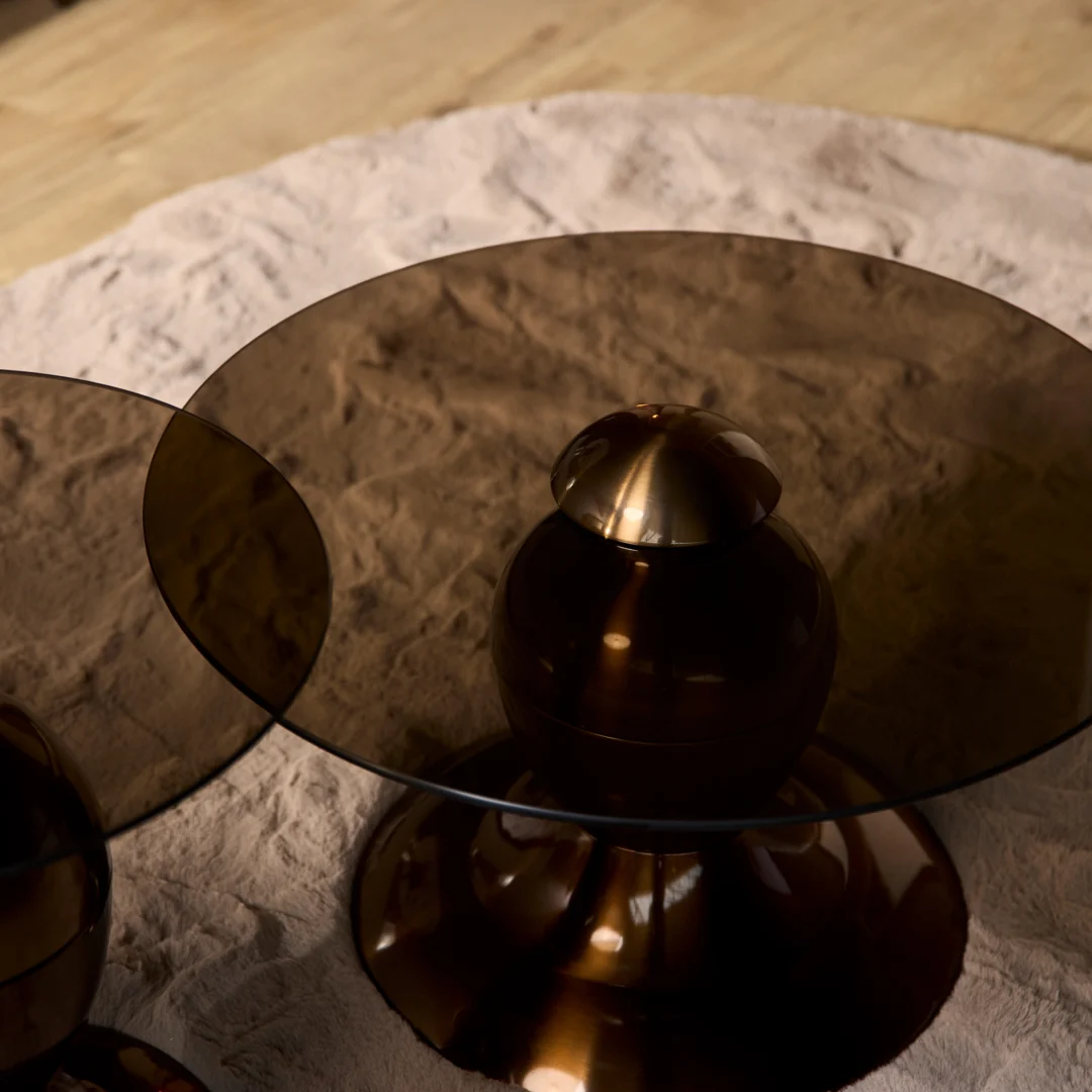 De Salontafel Industrieel, een ronde bronzen tafel met een donker glazen blad, staat op een lichtbeige vloerkleed met textuur op een houten vloer. Een andere soortgelijke Salontafel Industrieel is gedeeltelijk zichtbaar aan de linkerkant.