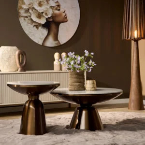 Een moderne woonkamer met twee Salontafel Design Tijdloos salontafels met minimalistisch design op een beige vloerkleed, een vaas met paarse bloemen, decoratieve objecten, een kast, wandkunstwerk van een vrouw met bloemen in haar haar en warme sfeerverlichting.