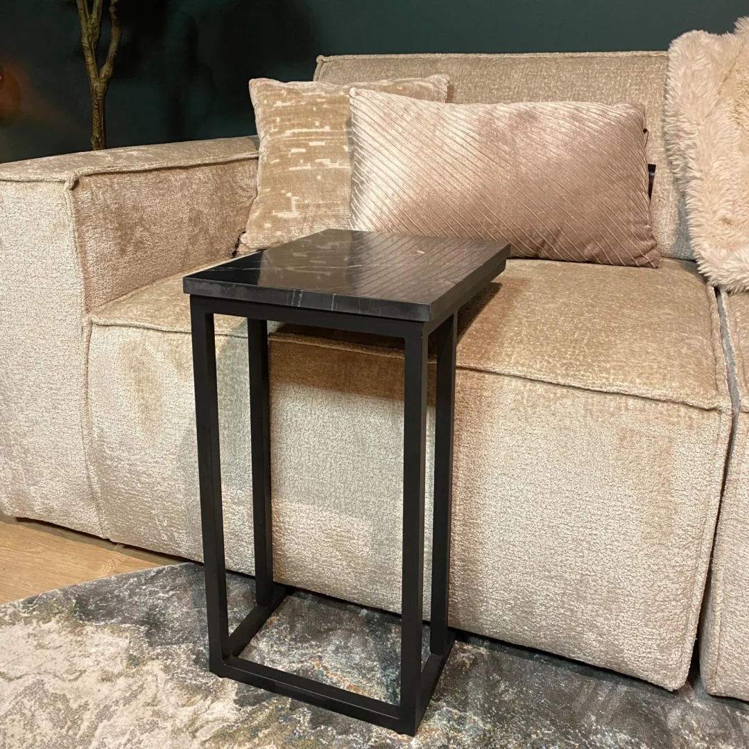 De Schuifregelaar Nova Zwart marmer, een klein zwart marmeren bijzettafeltje met een donker rechthoekig blad, staat voor een beige bank met gestructureerde kussens op een lichtgekleurd vloerkleed met patroon.