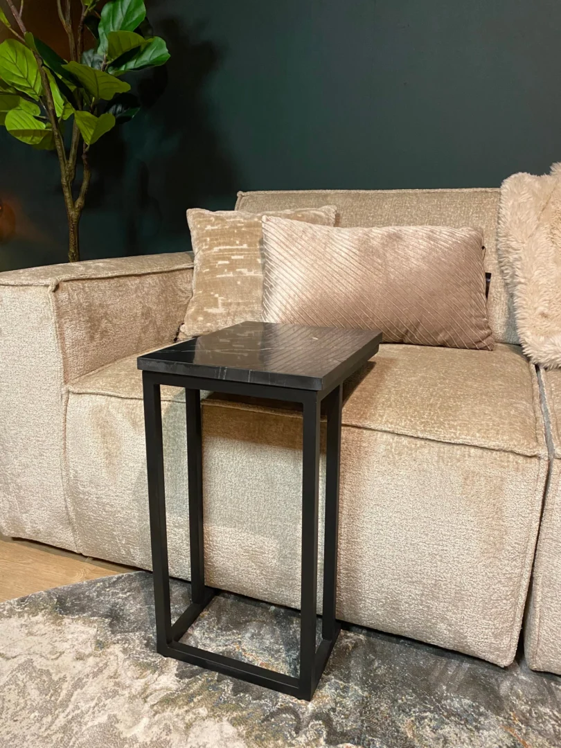 De Schuifregelaar Nova Zwart marmer, een compacte zwart marmeren bijzettafel met een glanzend oppervlak, staat voor een beige gestructureerde bank met pluche kussens. Op de achtergrond staat een groene plant en een vloerkleed met patroon.