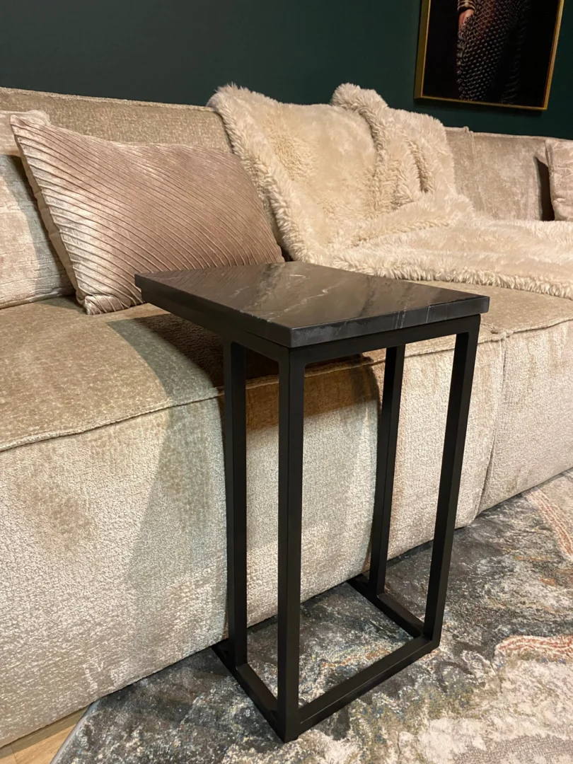 De Schuifregelaar Nova Zwart marmer, een compacte bijzettafel met een zwart metalen frame en een marmeren blad, staat voor een beige bank versierd met structuurkussens en een harige deken tegen een donkergroene muur.