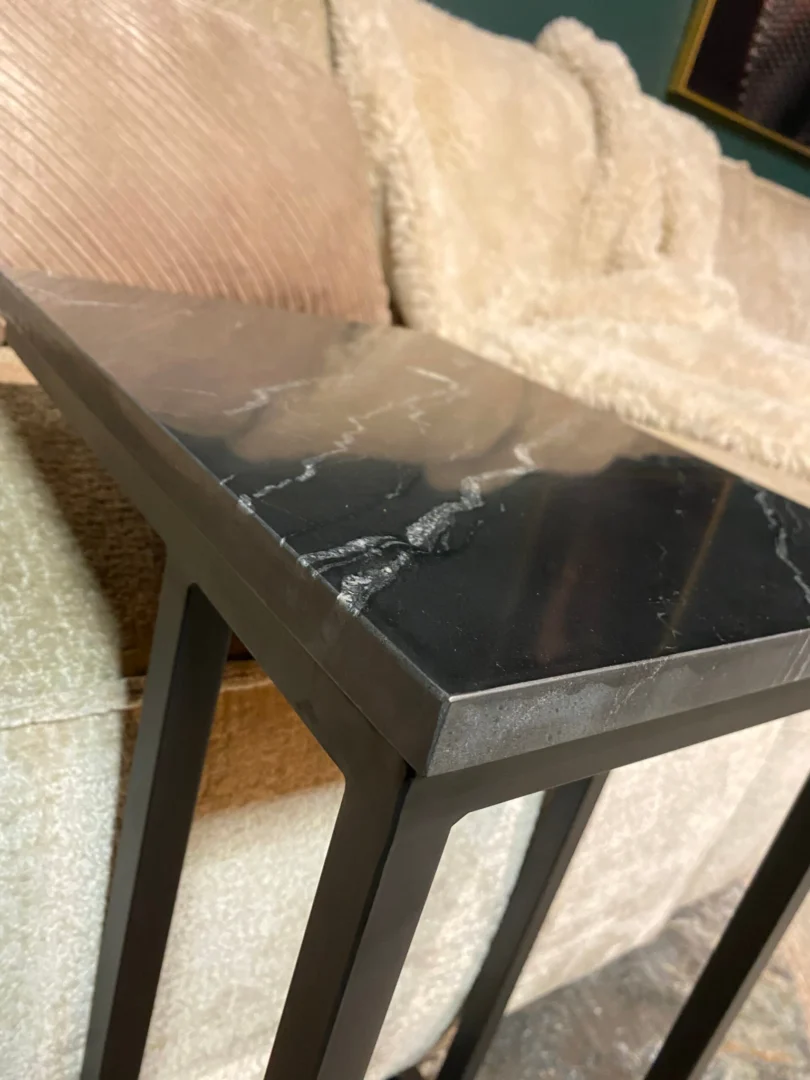 Een close-up van de Schuifregelaar Nova Zwart marmer bijzettafel met metalen poten, naast een beige gestructureerde bank versierd met lichtgekleurde pluche kussens en een knusse deken.