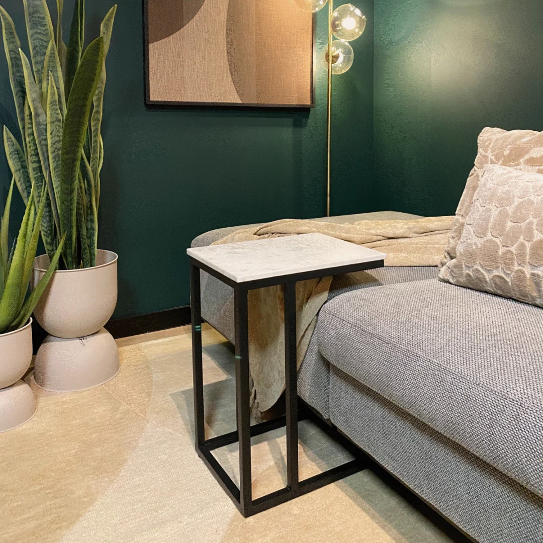 Een moderne woonkamer met een grijze bank, de Schuifregelaar Nova bijzettafel in wit marmer, een hoge groene potplant, een beige vloerkleed, structuurkussens en een gouden staande lamp tegen donkergroene muren.