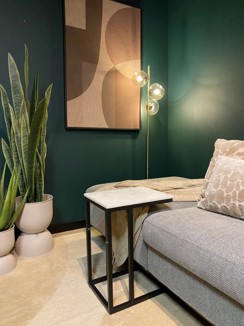 Een moderne woonkamer met een grijze bank, gedessineerde kussens, de Schuifregelaar Nova Wit marmer bijzettafel, een slangenplant in witte potten, een messing vloerlamp met glazen bollen en geometrische muurkunst op een donkergroene muur.